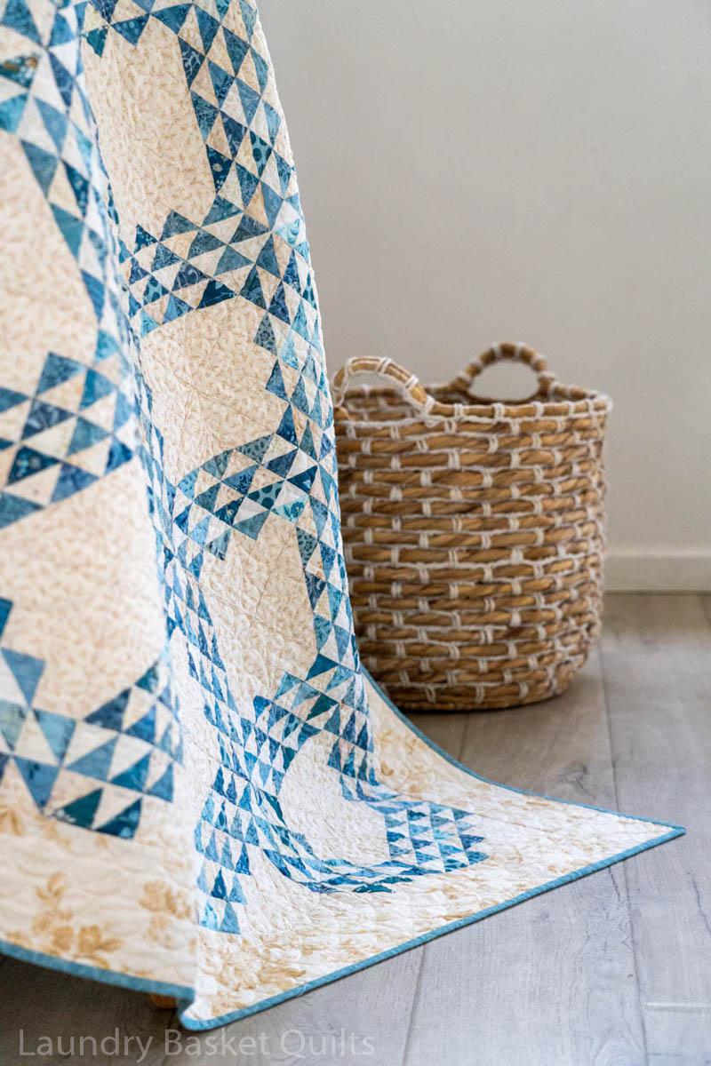 A blue and cream geometric patterned quilt draped over a woven basket on a light wood floor against a neutral wall.