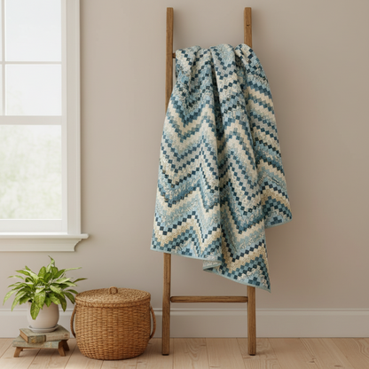 A crocheted throw blanket with a blue and cream chevron pattern is draped over a wooden ladder in a styled room setting with a window, potted plant, and wicker basket.