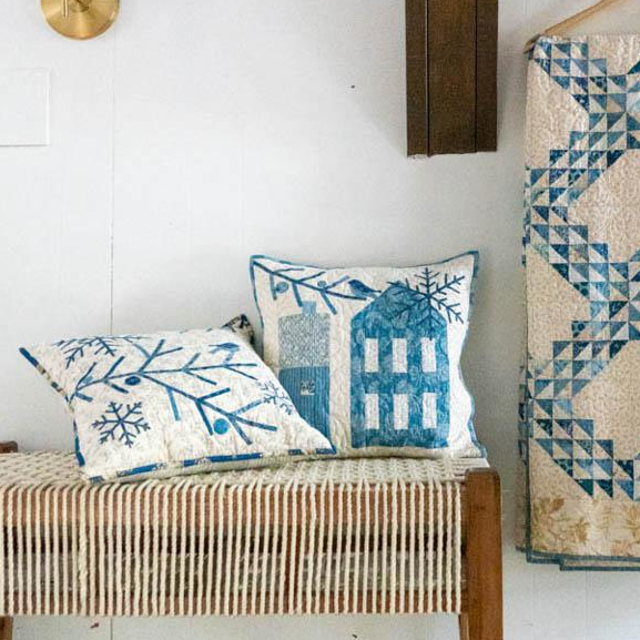 A styled interior scene showing a woven bench with two blue-and-white patterned throw pillows, alongside a matching quilted textile hanging on a hanger against a white paneled wall.
