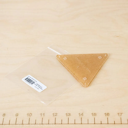 A triangular acrylic block with a clear plastic sleeve and a white barcode label, placed on a light wood surface with a ruler visible at the bottom.
