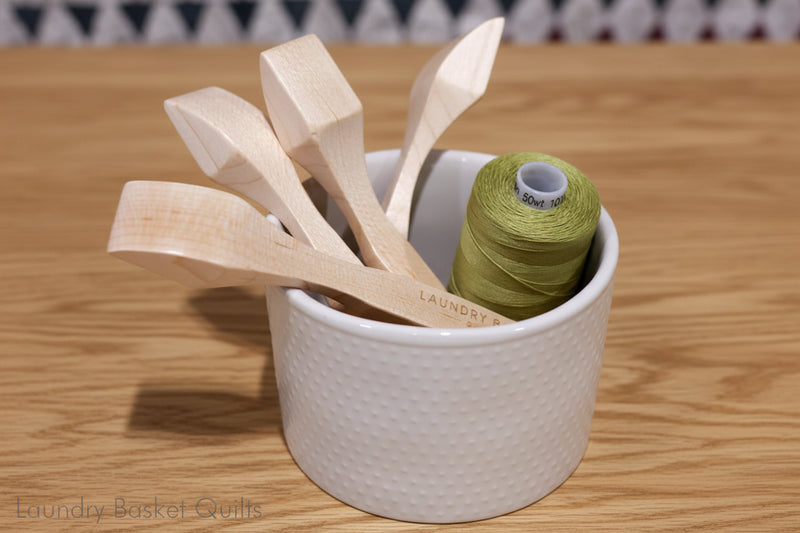 Wooden spoons and a spool of green thread in a white container labeled 'Laundry Basket Quilts'.