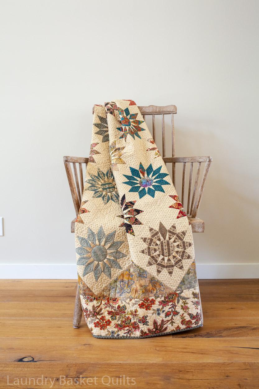 A vintage-style quilted blanket with star and floral motifs draped over a wooden spindle chair in a room with a light beige wall and hardwood floor.