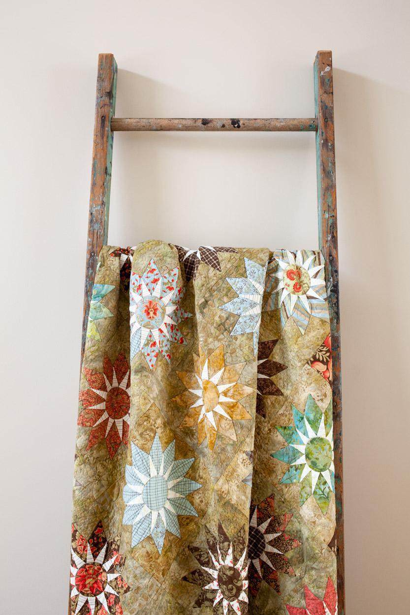 A vintage-style rustic wooden ladder draped with a multi-colored quilt featuring starburst and floral patterns in beige, blue, red, and green, set against a plain light beige wall.