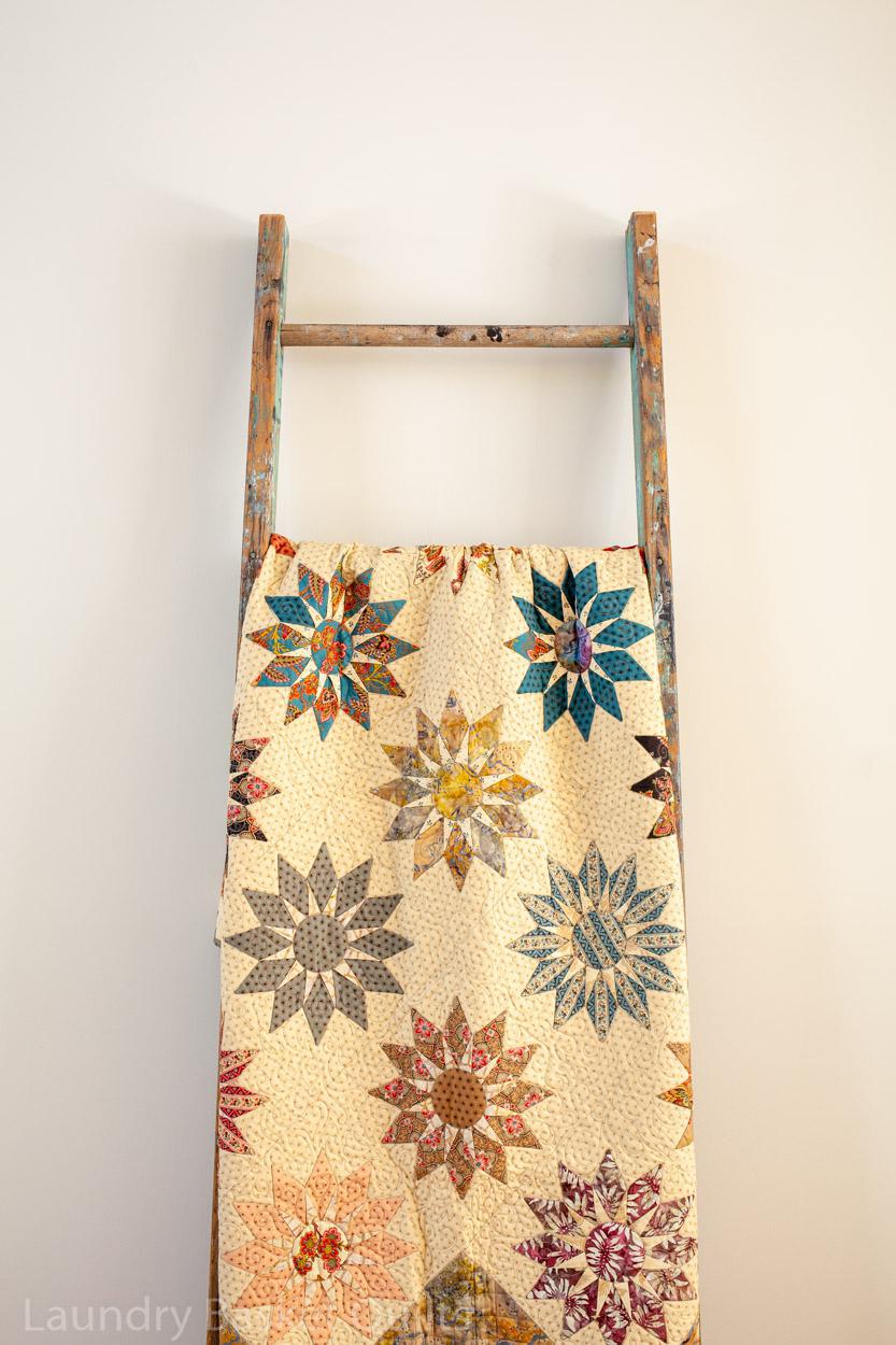A vintage-style quilted fabric panel with a colorful starburst floral pattern, draped over a rustic wooden clothes ladder against a plain off-white wall.