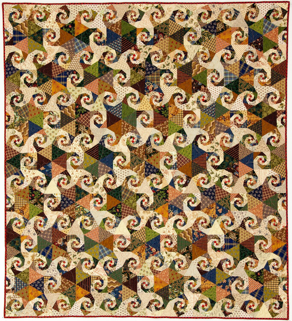 A vintage-style patchwork quilt featuring a repeating swirl pattern made from many small fabric triangles in earthy tones of brown, beige, blue, green, orange, and cream, finished with a red border.