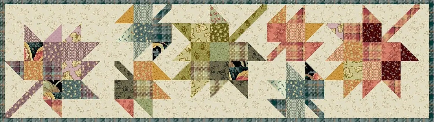 A quilted fabric banner featuring five stylized starburst or pinwheel motifs in various floral and plaid patterns, set against a cream background with a green plaid border.