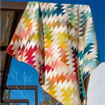 A colorful quilted blanket with a rainbow geometric pattern draped over a wooden structure against a blue sky, 