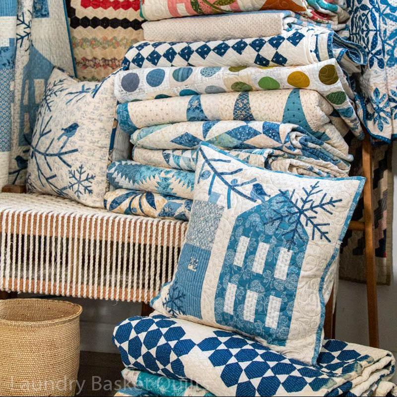 A collection of blue and white patterned quilts and pillows displayed on a bench and stacked in a bundle, featuring various geometric and floral designs.