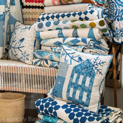 A collection of blue and white patterned quilts and pillows displayed on a bench and stacked in a bundle, featuring various geometric and floral designs.