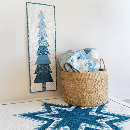 A woven natural-fiber storage basket filled with folded blue and white patterned textiles, placed beside a tall vertical quilted tree-patterned wall hanging on a light wall above a blue star rug.