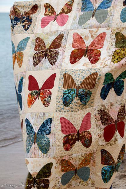 A close-up of a quilted fabric panel featuring a patchwork of colorful butterfly appliqués in red, blue, green, brown, and pink on a cream background, with a soft-focus beach and water background.