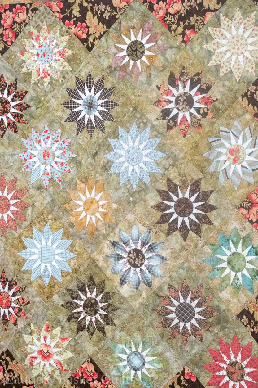A close-up of a quilted fabric featuring a repeating pattern of multi-colored starburst and floral quilt blocks on a textured beige background.