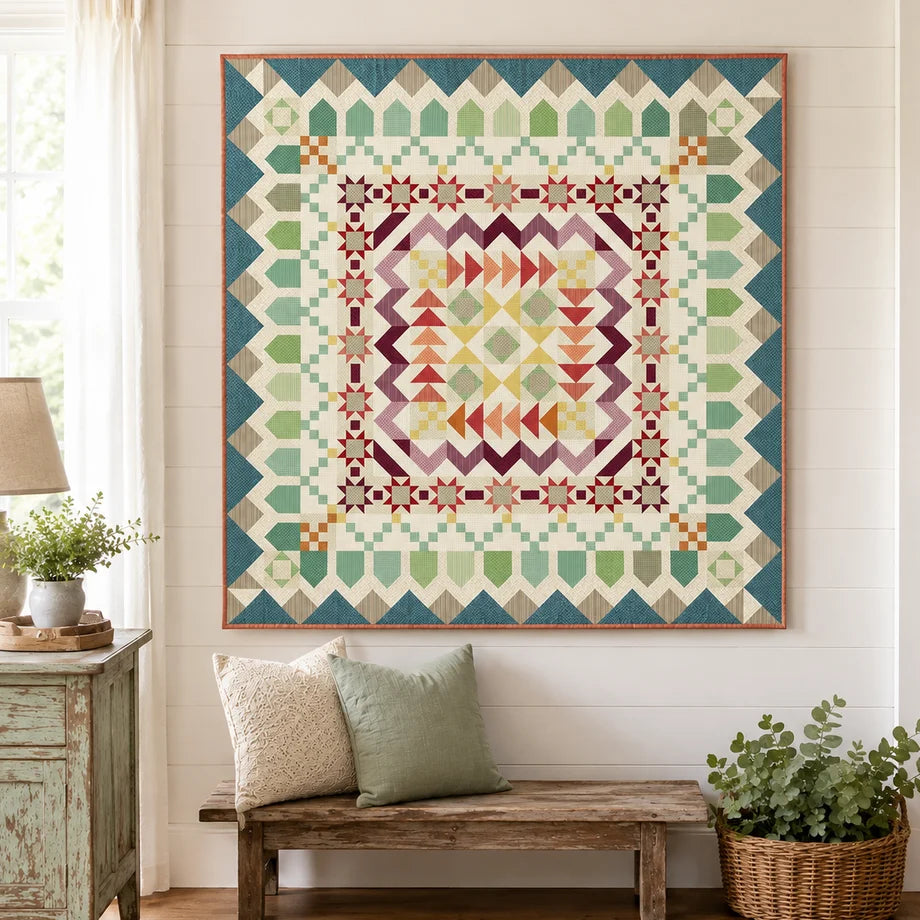 A large framed geometric quilt hanging on a white paneled wall above a rustic wooden bench with two pillows in a bright, styled room.
